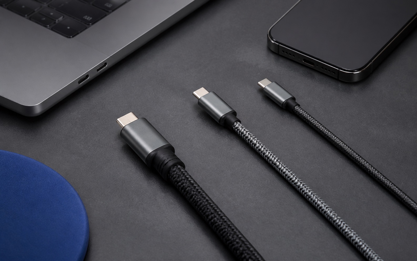 Three unbranded braided USB-C cables beside a laptop and smartphone on a graphite desk